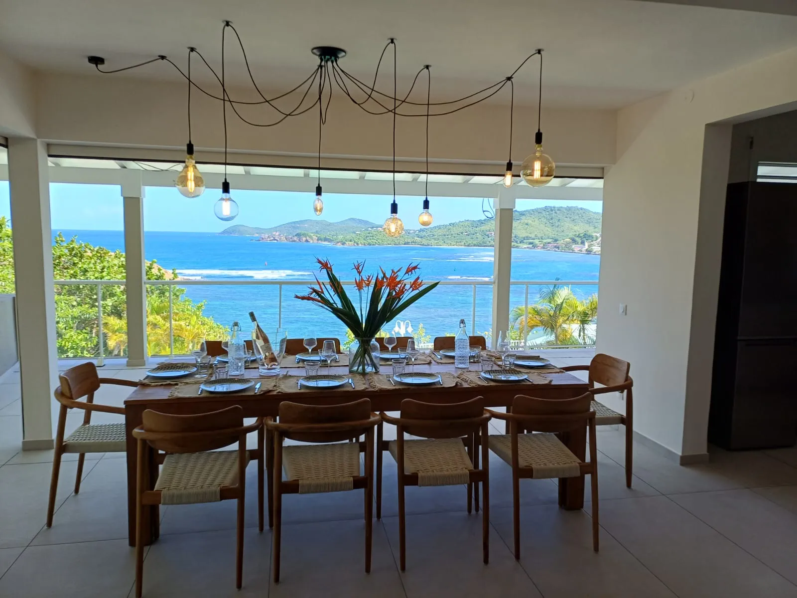Grande table dressée pour dix sous une suspension d'ampoules, ouverte sur la vue mer panoramique de Tartane.